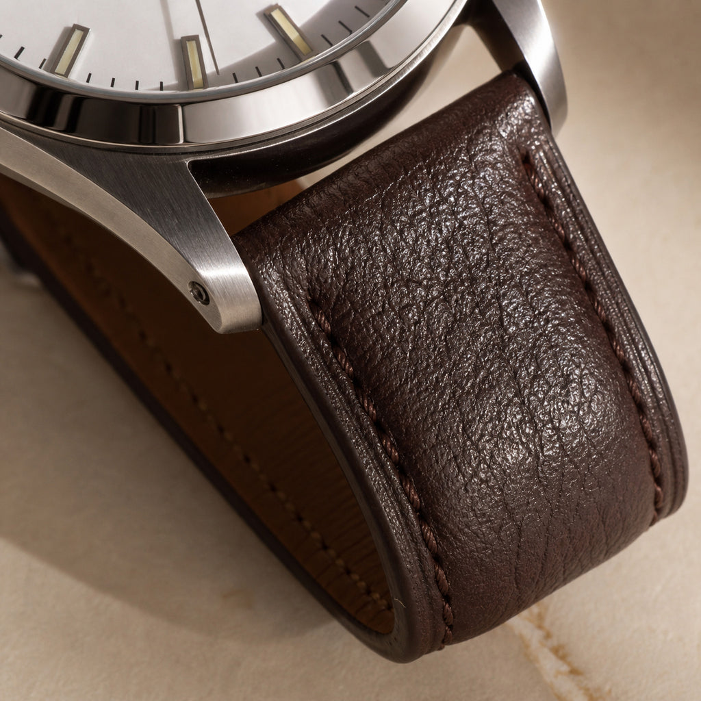 Quarante-et-Un Blanc — macro de la jonction entre la corne acier brossée et le bracelet cuir brun foncé grainé, surpiqûre ton sur ton, tranche de cadran blanc visible — Maison Noirmont