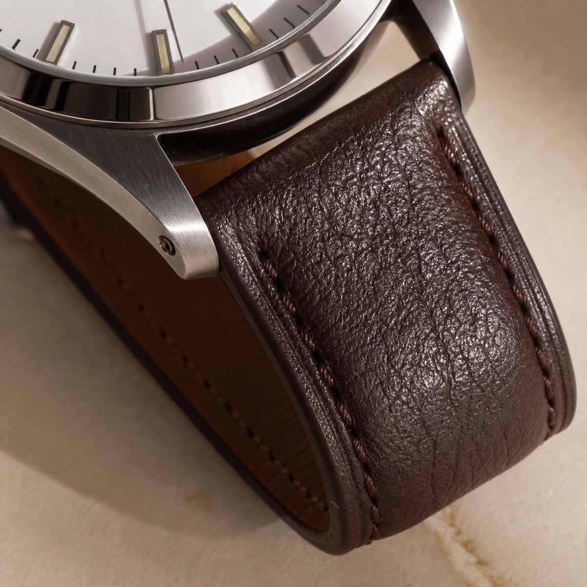 Quarante-et-Un Blanc — macro de la jonction entre la corne acier brossée et le bracelet cuir brun foncé grainé, surpiqûre ton sur ton, tranche de cadran blanc visible — Maison Noirmont