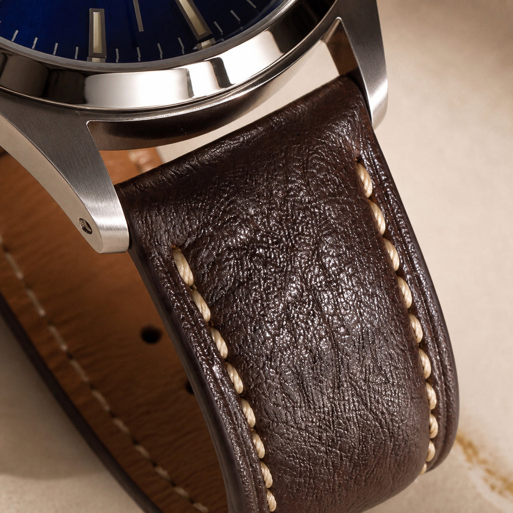 Quarante-et-Un Bleu — macro de la jonction entre la corne acier brossée et le bracelet cuir brun foncé à surpiqûres beige, tranche de cadran bleu roi visible — Maison Noirmont
