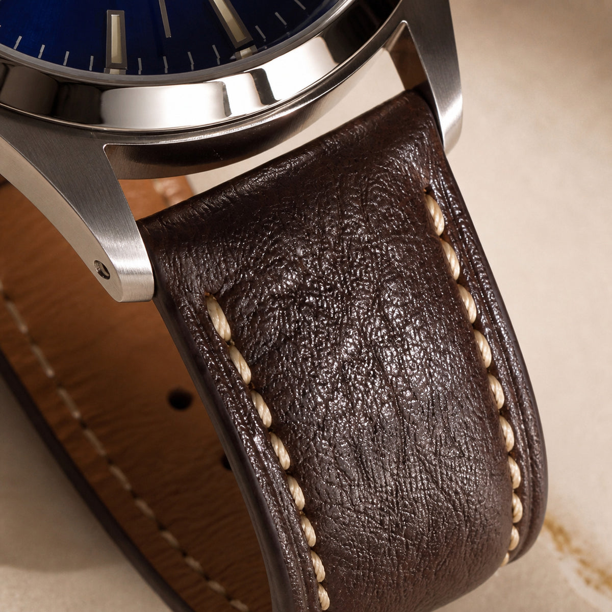 Quarante-et-Un Bleu — macro de la jonction entre la corne acier brossée et le bracelet cuir brun foncé à surpiqûres beige, tranche de cadran bleu roi visible — Maison Noirmont