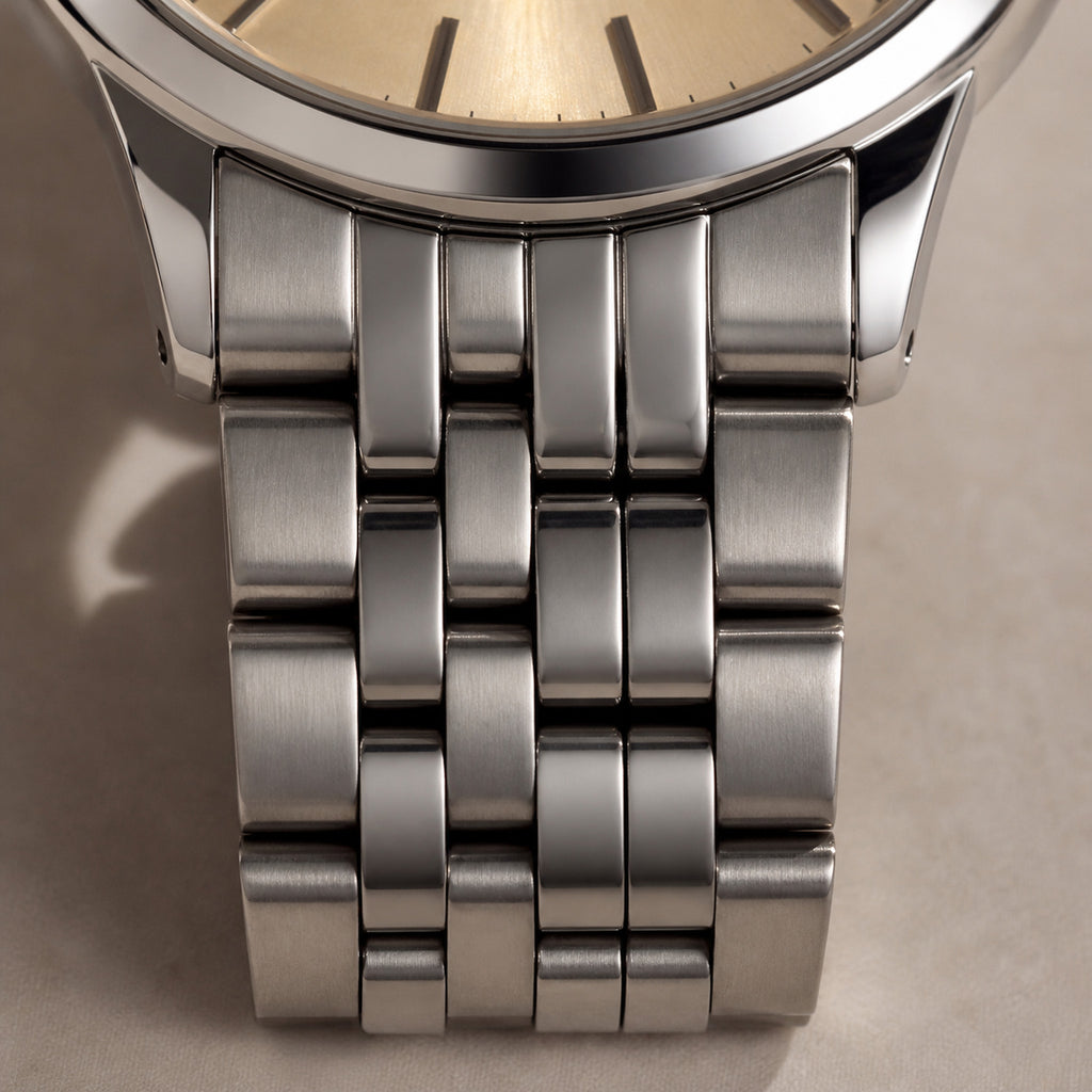 Trente-Six Doré — macro frontale de la jonction boîtier-bracelet : cadran champagne soleillé à index acier en haut de cadre, boîtier et bracelet cinq rangs en acier — Maison Noirmont