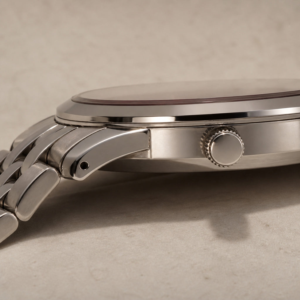Trente-Six Rouge — profil rapproché : boîtier acier fin, couronne cannelée à sommet lisse, tranche de cadran rouge cramoisi sous le verre et première articulation du bracelet cinq rangs — Maison Noirmont
